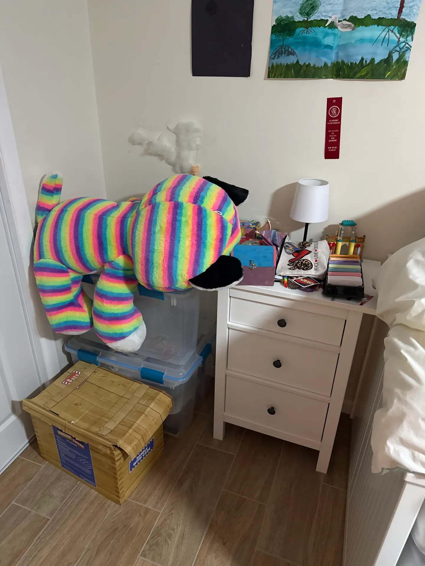 A child’s bedroom corner with a nightstand surrounded by a plush toy, boxes, books, and small items before organizing