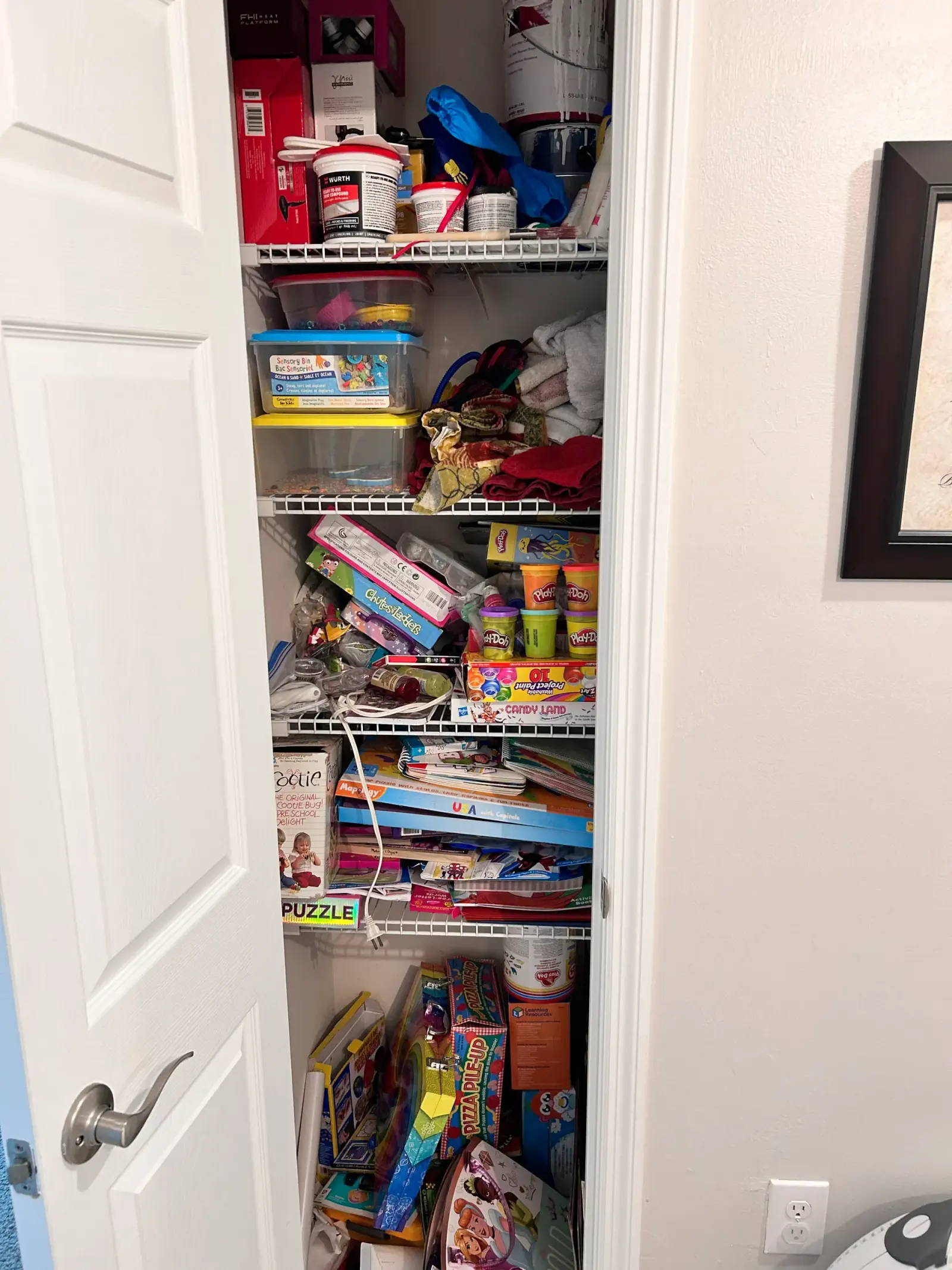A craft and play closet with games, art supplies, fabric, and learning materials arranged across the shelves before organizing