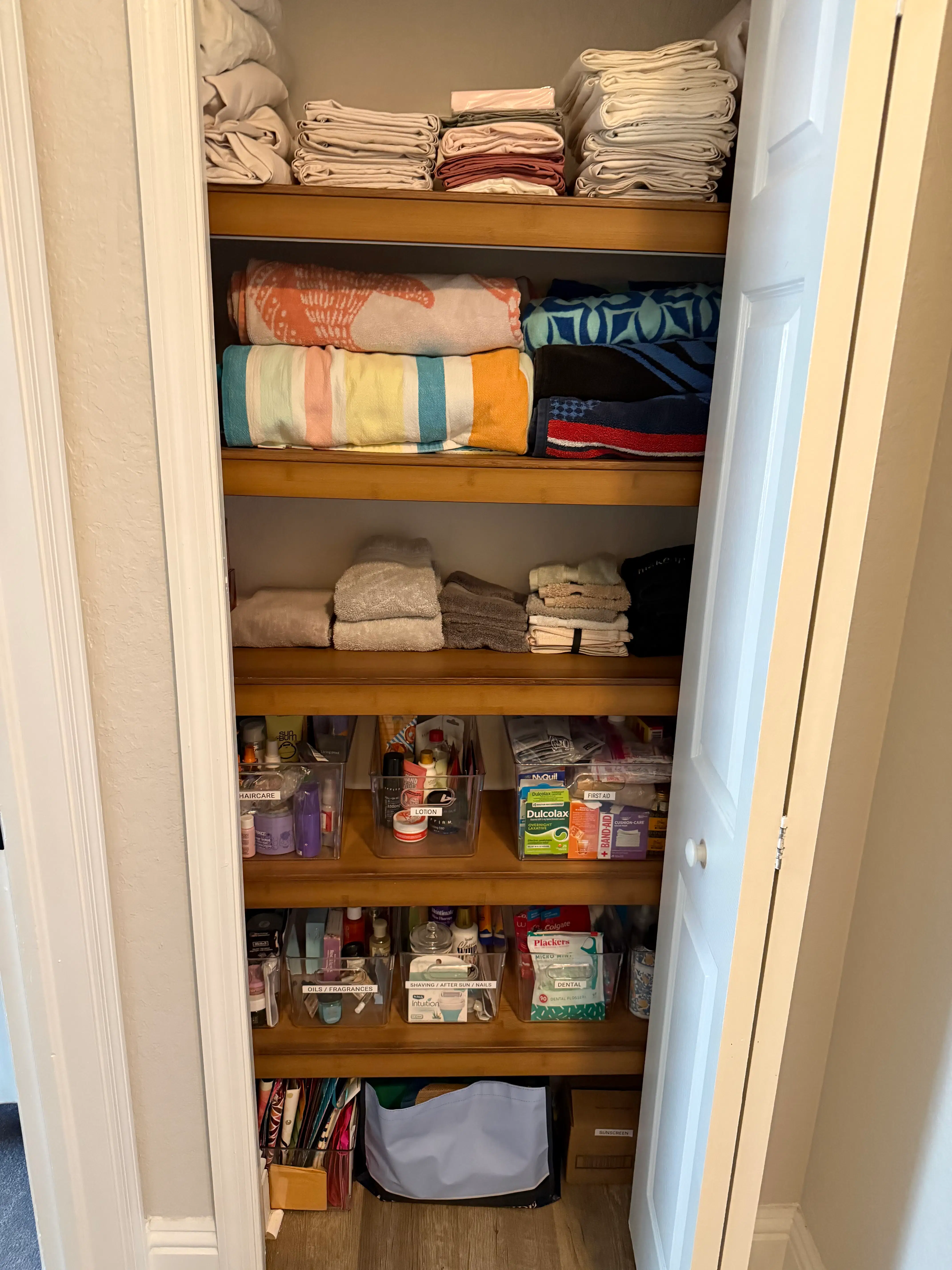 An organised linen closet with folded linens and towels stacked on the upper wooden shelves and clear acrylic bins of categorised haircare, dental, first-aid, and toiletry supplies on the lower shelves, labelled so everything is easy to find